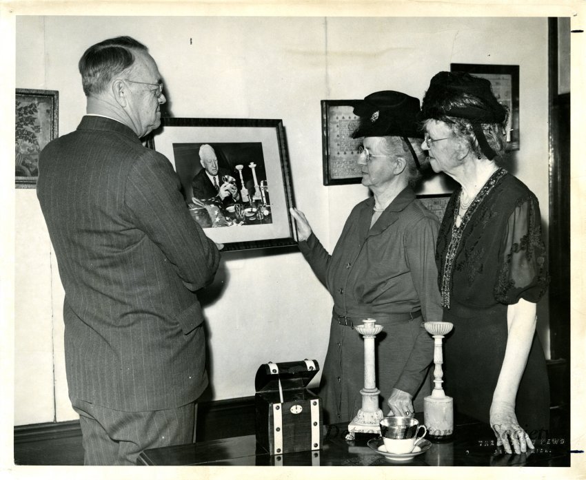 Black and white photo of J. Bell Moran, Gracie Krum and Mrs. Arthur S. Hampton at the Detroit Historical Museum. Black and white photographic print of (left to right) J. Bell Moran; Gracie B. Krum; and Mrs. Arthur S. Hampton, viewing photographs, and artifacts inside the Detroit Historical Museum, 1949.
