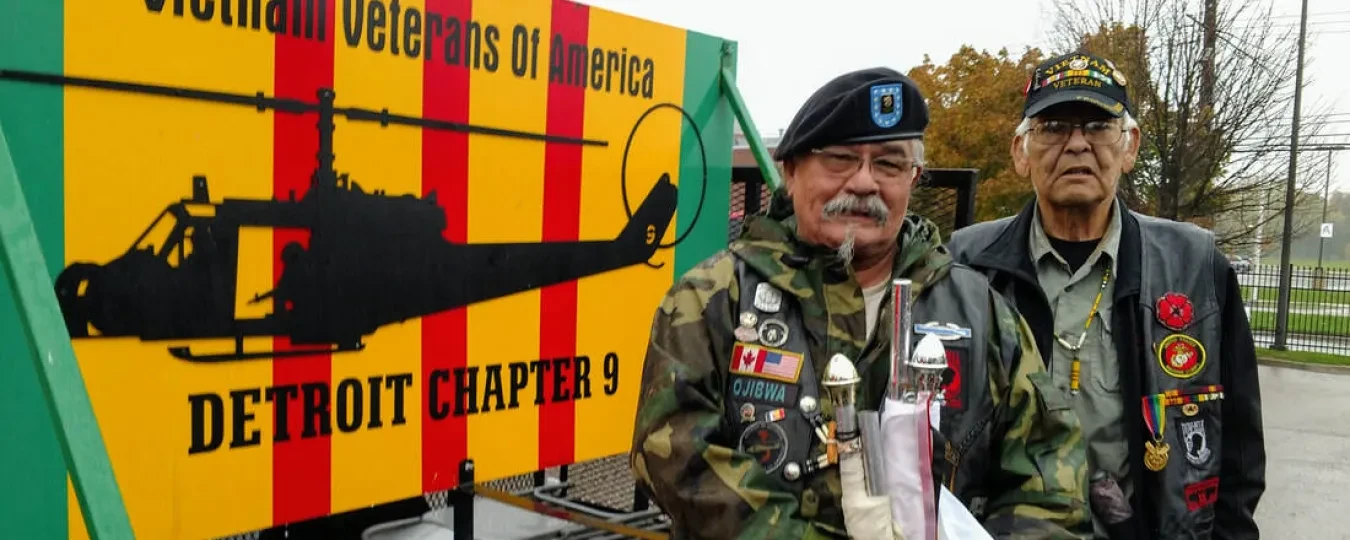 Two elderly veterans in decorated jackets stand beside a Vietnam Veterans of America sign outdoors.