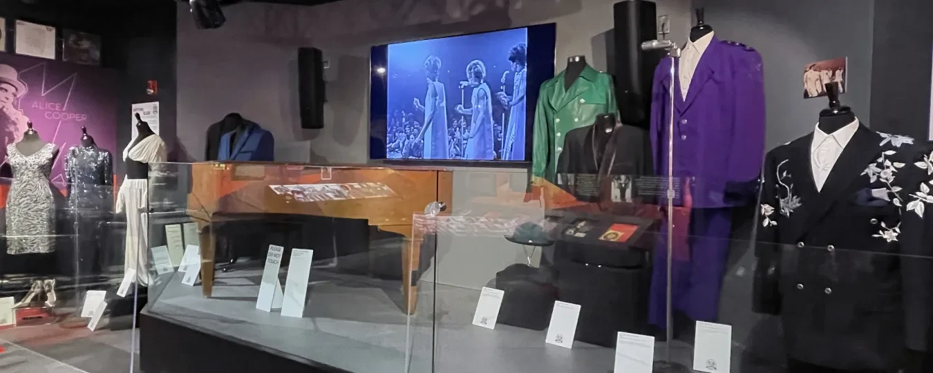 Museum display of vintage mannequins in colorful suits behind glass, with a screen showing historical footage.