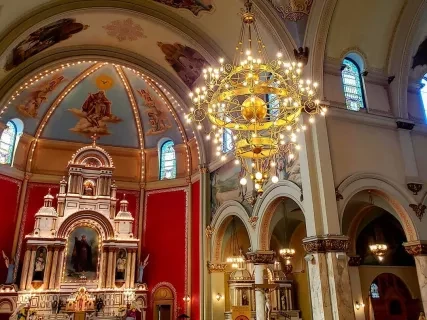 Interior of ornate catholic church with lighted crystal chandelier hanging from the painted ceiling and an altar in front of a red wall.