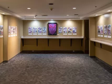 Art gallery with framed pictures on beige walls, focused lighting, and carpeted floor. Central artwork features a large purple shape.