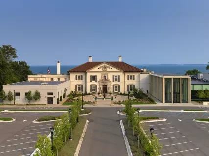 Elegant mansion with a terracotta roof, surrounded by manicured gardens and parking, set against a clear blue sky.