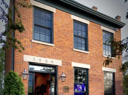 Brick building with a black door, steps, and plants. Large windows reflect sky, surrounded by trees and greenery.