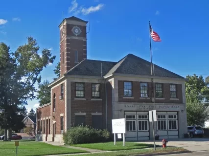 Historic brick fire station with a clock tower, flag, and garage doors. Clear blue sky background.
