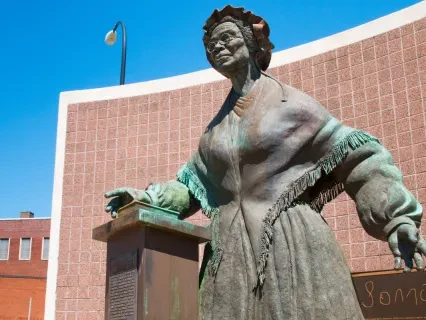 Bronze statue of a woman in a flowing dress against a blue sky, with a lamppost and brick building in the background.