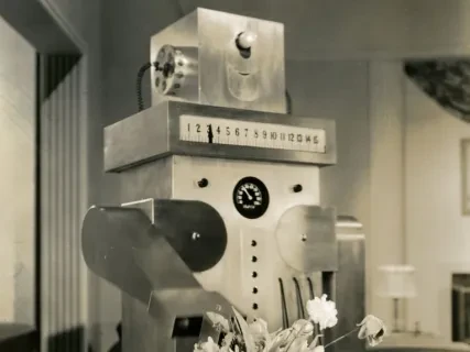 Retro robot in a room, positioned behind a vase of flowers on a table, with an award-like figurine to the right.
