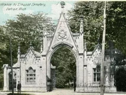 Gothic archway entrance to a cemetery, surrounded by trees and a person standing near the gate.