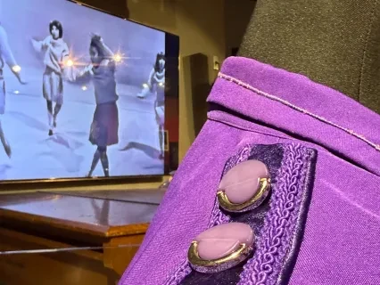 Purple jacket with ornate buttons on a mannequin, TV screen in the background, showing dancers.