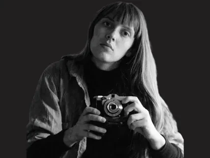 Black and white photo of a person with long hair holding a camera against a dark background.