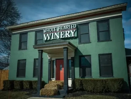 Two-story green building with a sign reading "Winery," red door, and hay bales in front, under a cloudy sky.