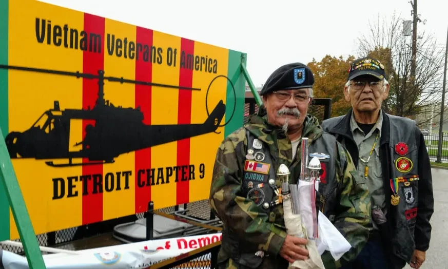 Two elderly veterans in decorated jackets stand beside a Vietnam Veterans of America sign outdoors.
