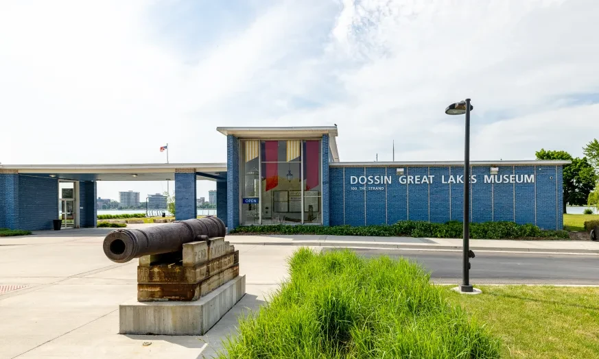 Cannon displayed outside a modern museum with a blue facade and large windows, surrounded by greenery.