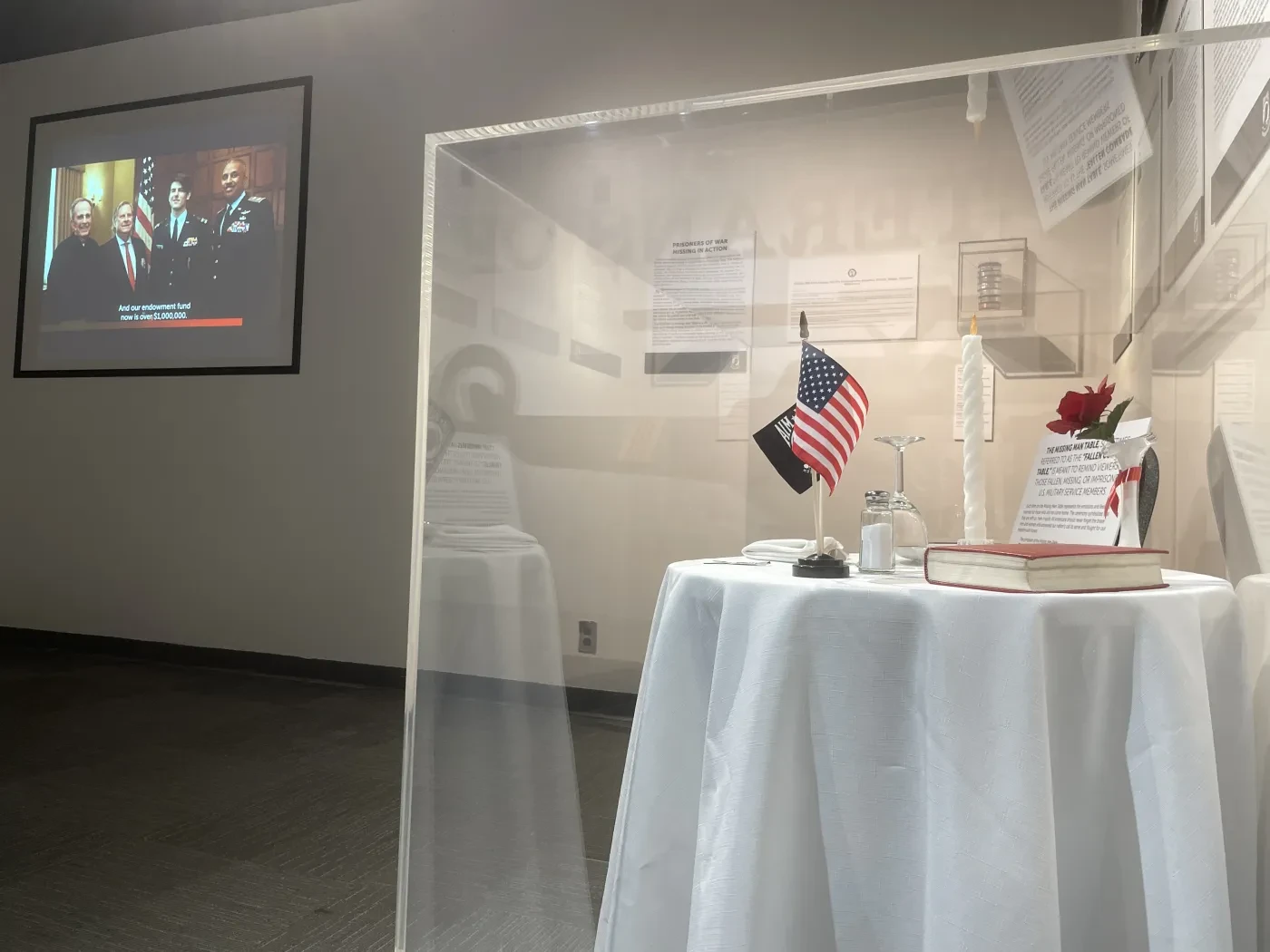 Table with American flag under glass, documents, and a framed TV screen displaying a news broadcast in a dimly lit room.