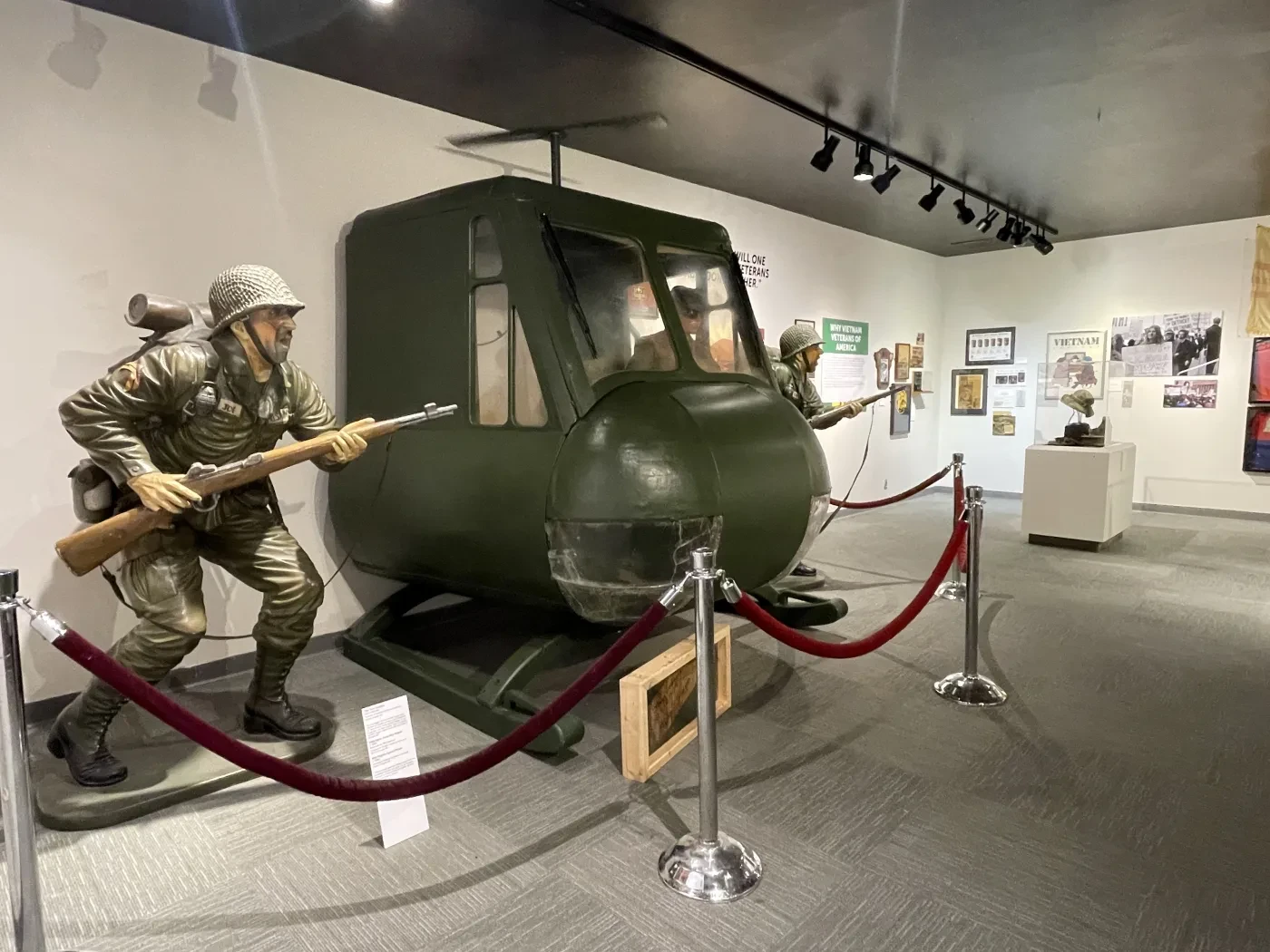 Museum display of a vintage helicopter cockpit with soldier mannequins, surrounded by red ropes.
