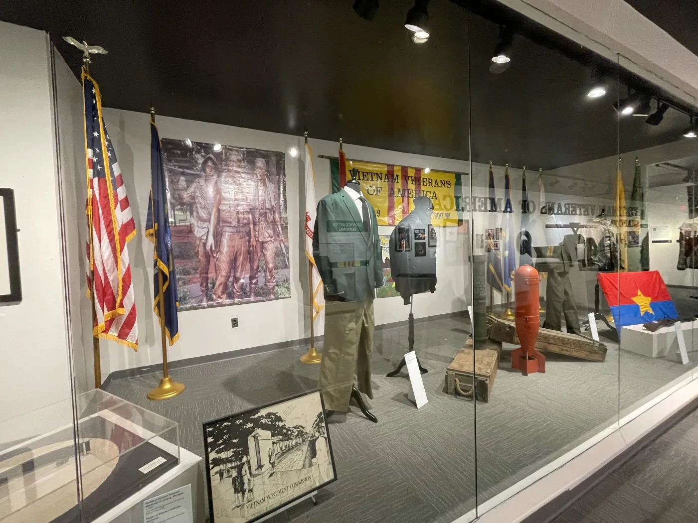 Museum exhibit with military uniforms, flags, and historical artifacts behind glass display.