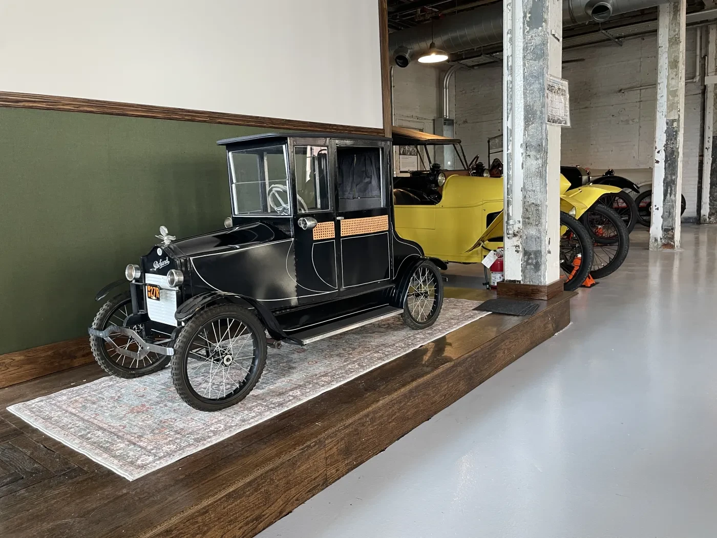 Vintage cars displayed on a raised platform in a museum, including a black vehicle with wooden accents and a yellow car behind.