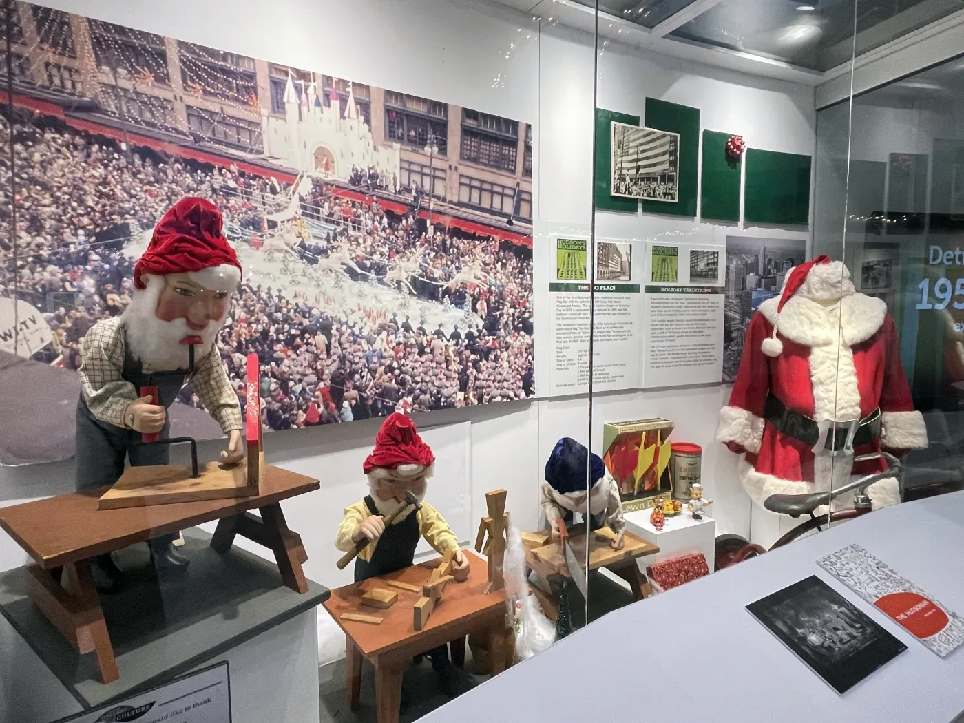 Elves in red hats and Santa in a Christmas display with toy workshop, under museum lighting.