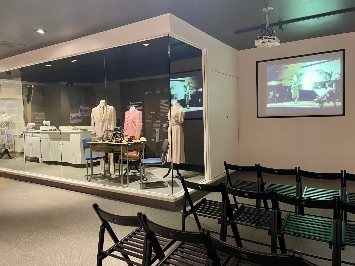 Mannequins in vintage clothing displayed next to a retro office setup; chairs face a projector screen displaying a presentation.
