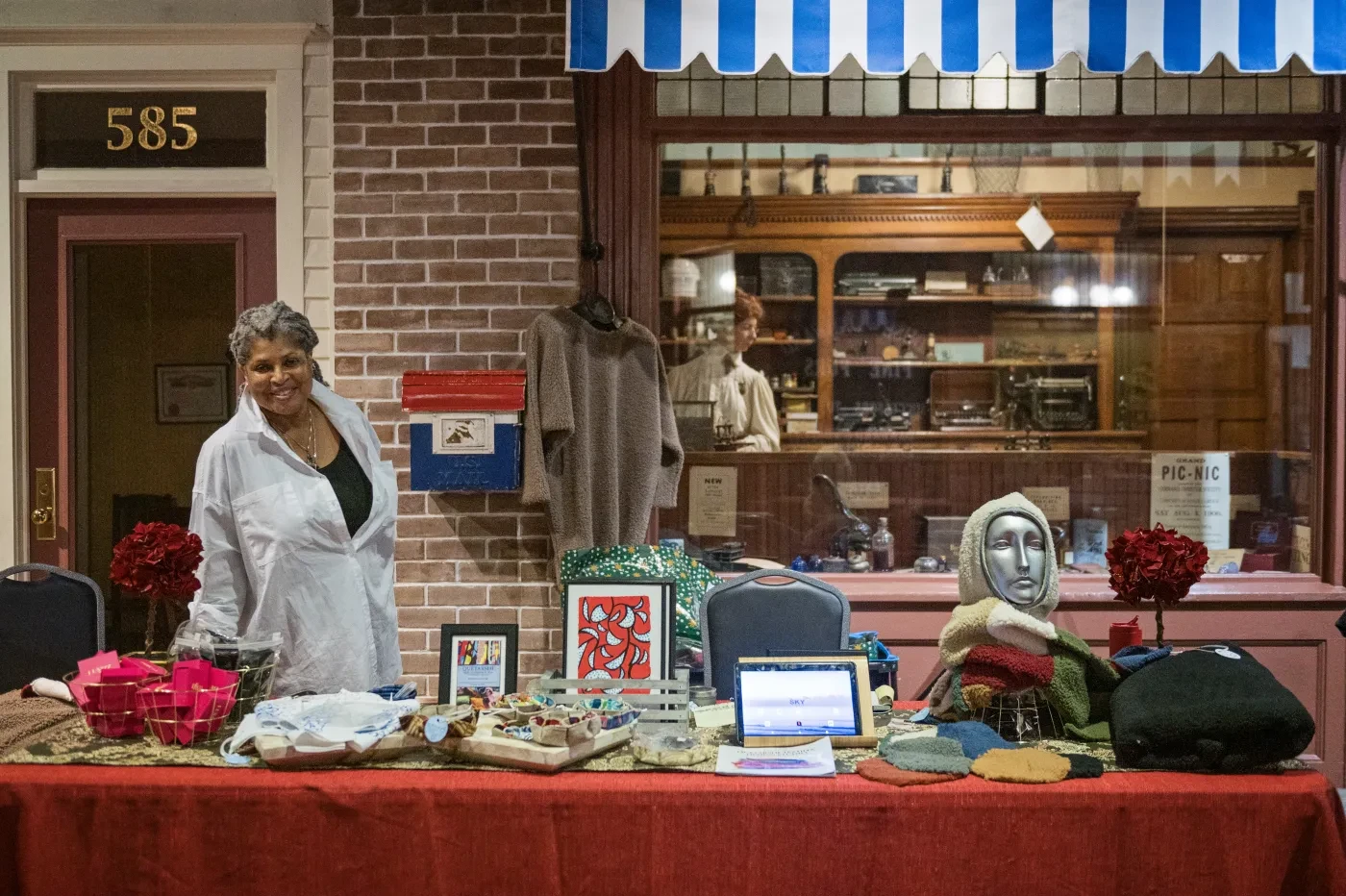 A smiling woman in a white coat at a craft table with handmade items, against a brick storefront with a blue-striped awning.