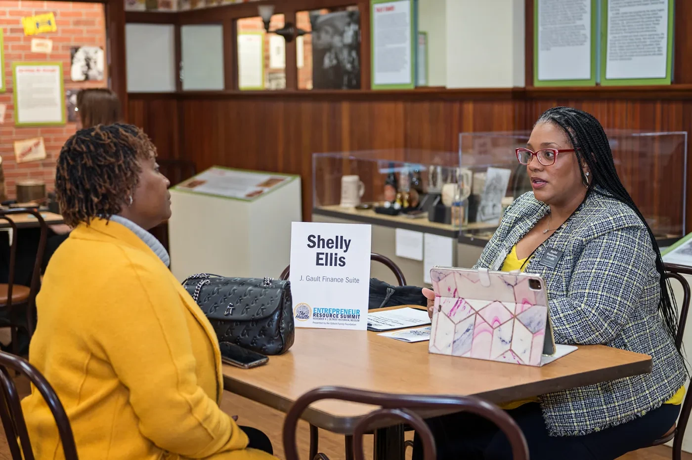 Two women sit at a table with a sign reading "Shelly Ellis." One is taking notes on a tablet.