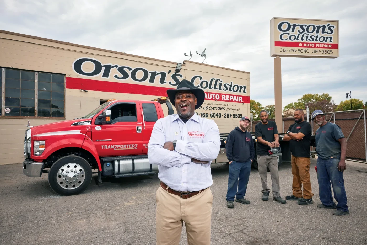 Man smiling in front of red tow truck and building with Orson's Collision sign; four people stand chatting nearby.