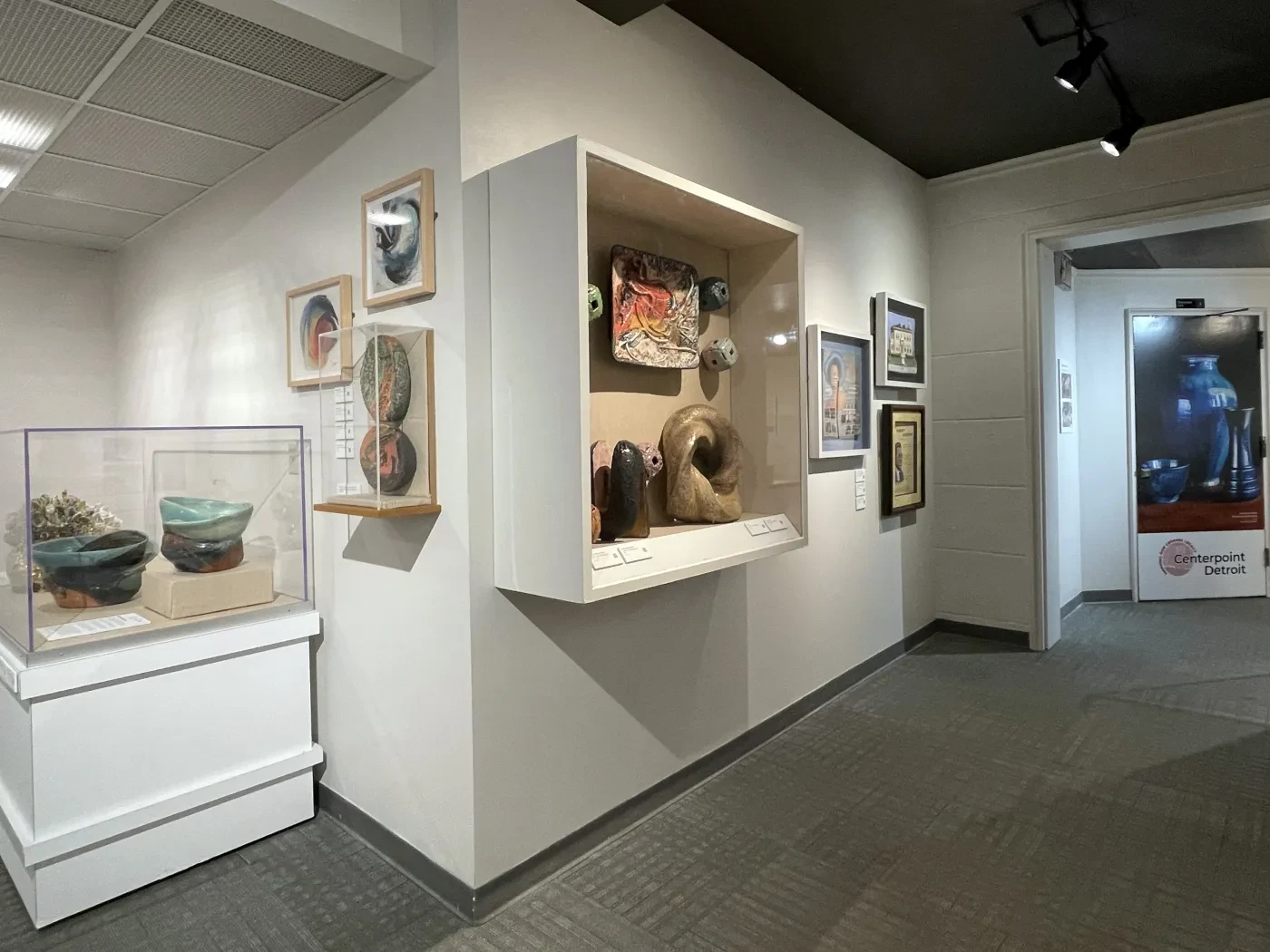 Museum hallway with art displayed on walls and in a glass case, featuring ceramics and framed pieces.