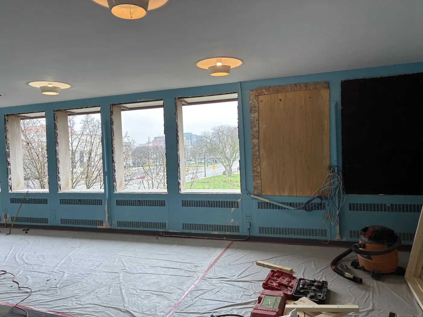 Room under renovation with large windows, light blue walls, construction materials, and tools scattered on the floor.