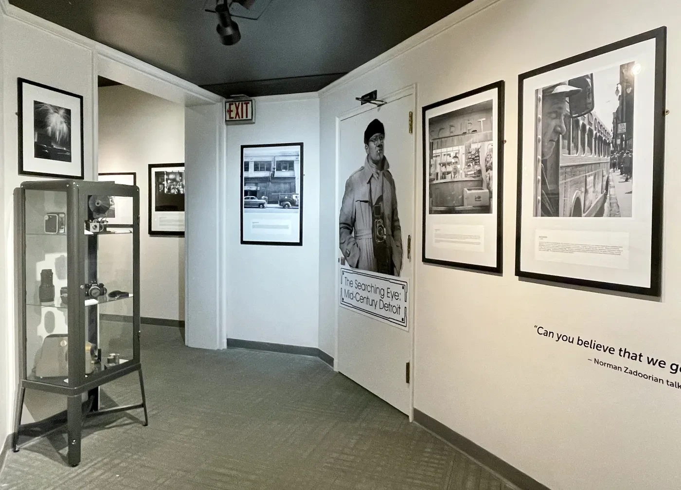 Museum hallway with black-and-white photographs on white walls, a clear display case to the left, and a dark ceiling.