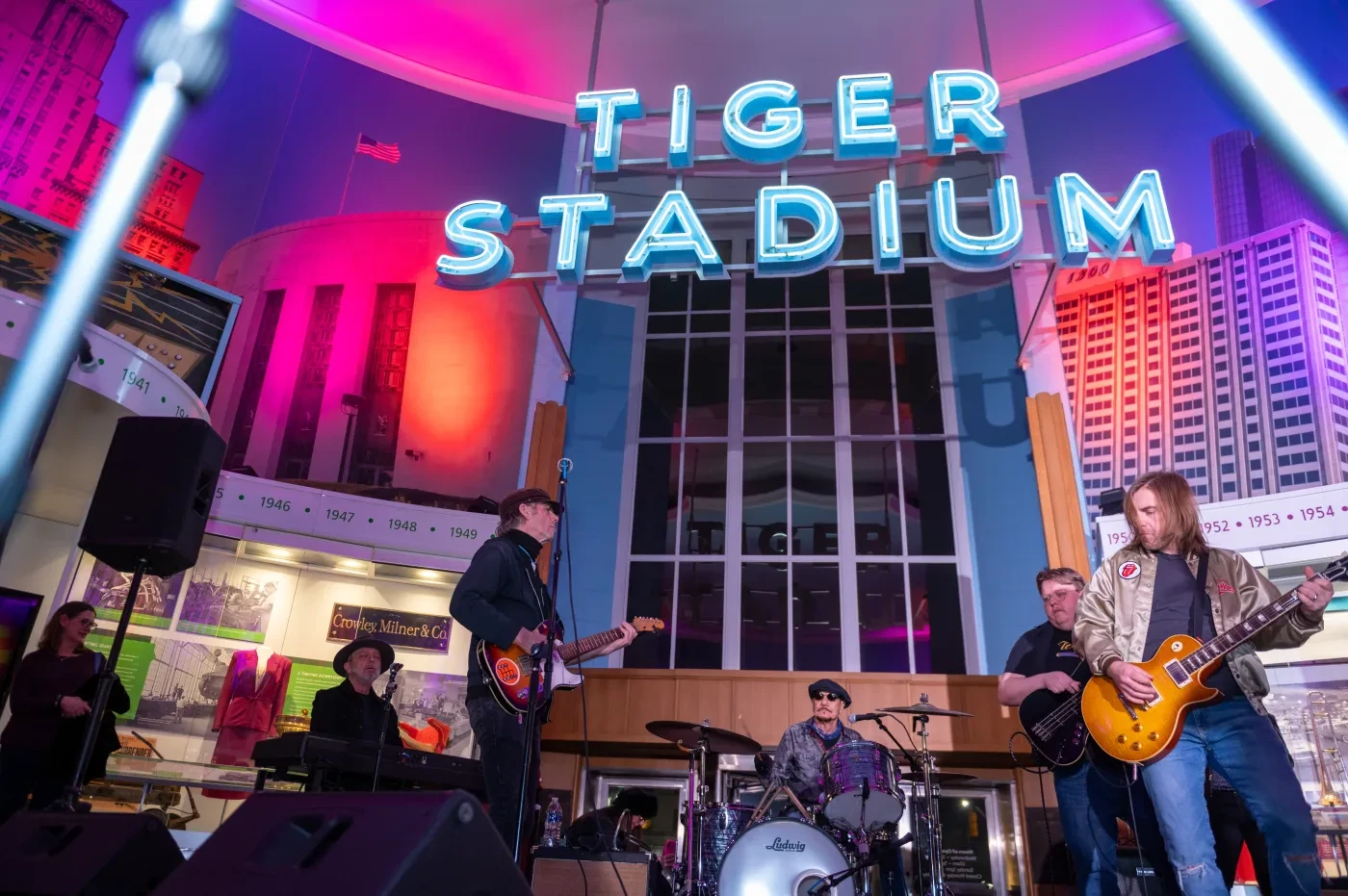 Band performing on stage under neon lights reading "Tiger Stadium," with vibrant pink and blue background.