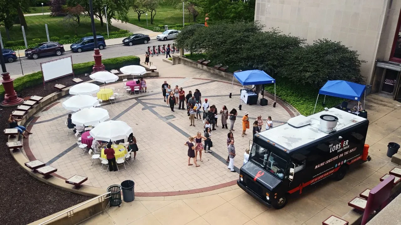 Outdoor gathering with people lining up at a food truck, tables with umbrellas, and surrounding greenery.