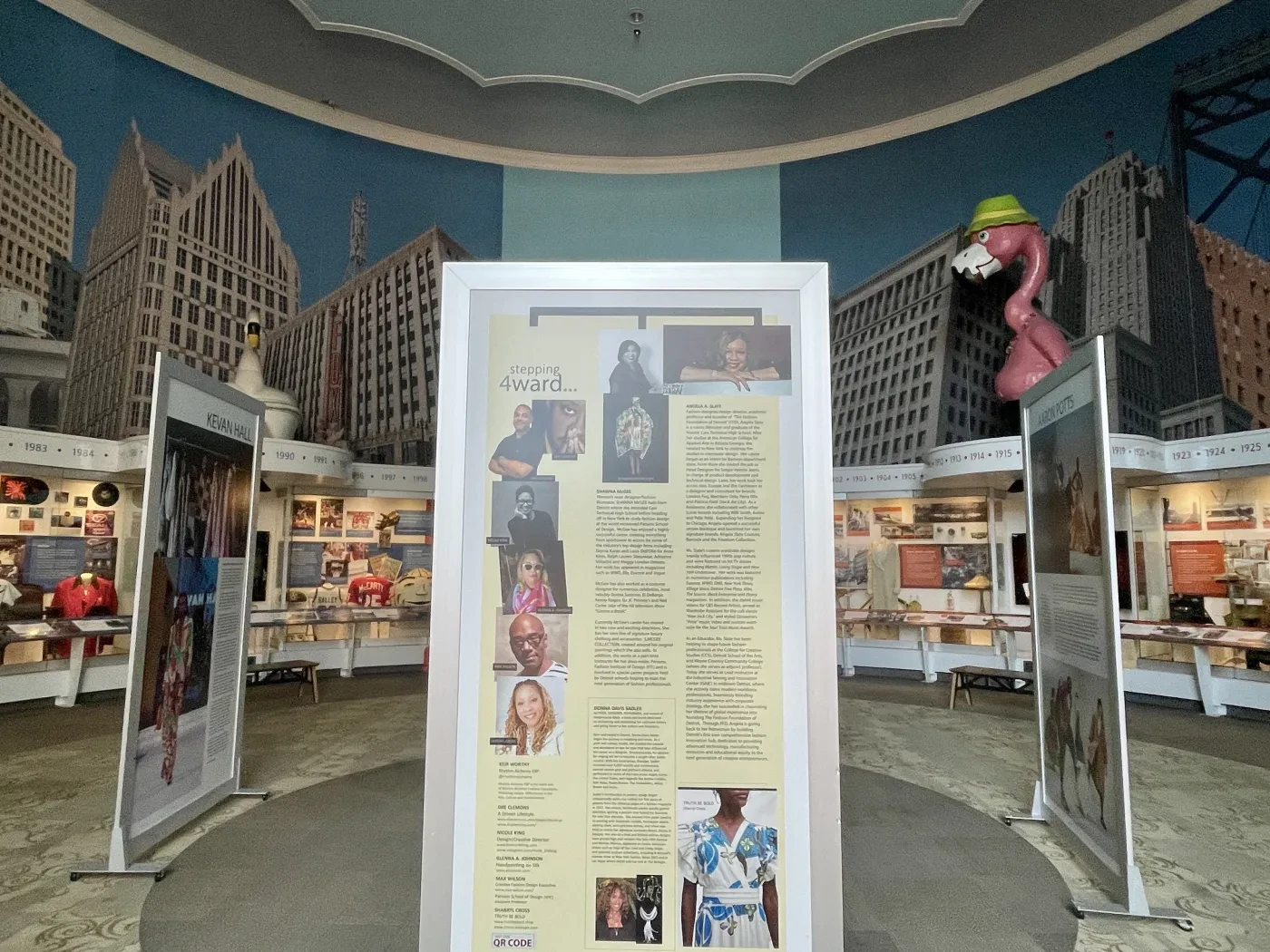 Exhibit room with cityscape mural, large display panels, and a giant flamingo statue wearing a green hat.