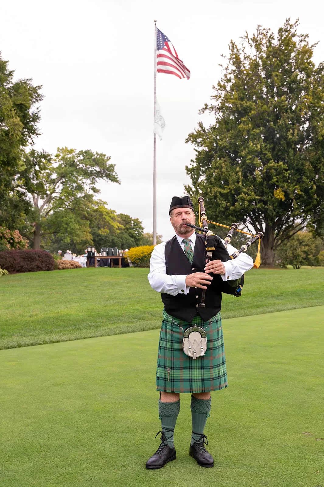 Man in a kilt playing bagpipes on a grassy field with trees and an American flag in the background.