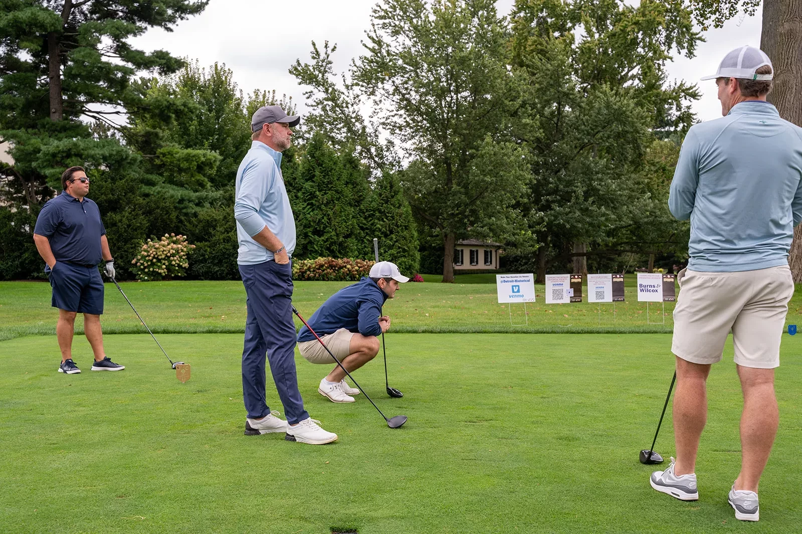 Four men on a golf course, one crouching with a club, surrounded by trees and signs in the background.