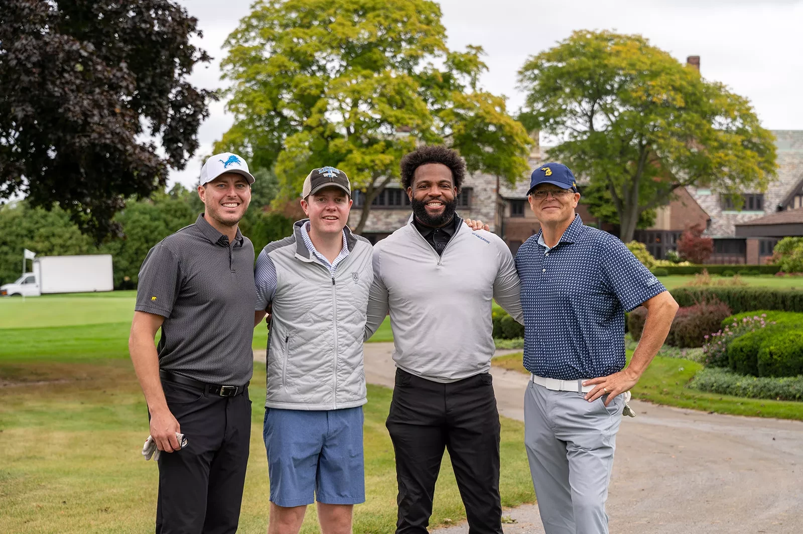 Four men smiling on a golf course, wearing casual golf attire, with trees and a house in the background.