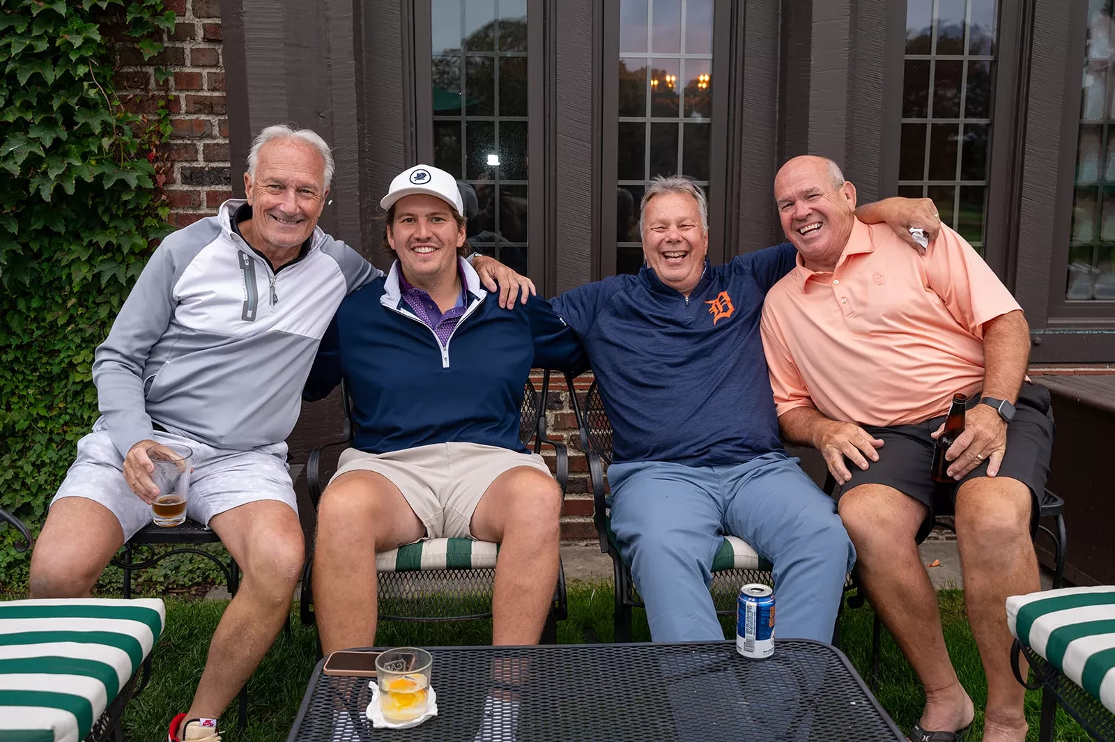 Four smiling men sitting together on patio chairs, with arms around each other, in front of a brick building.
