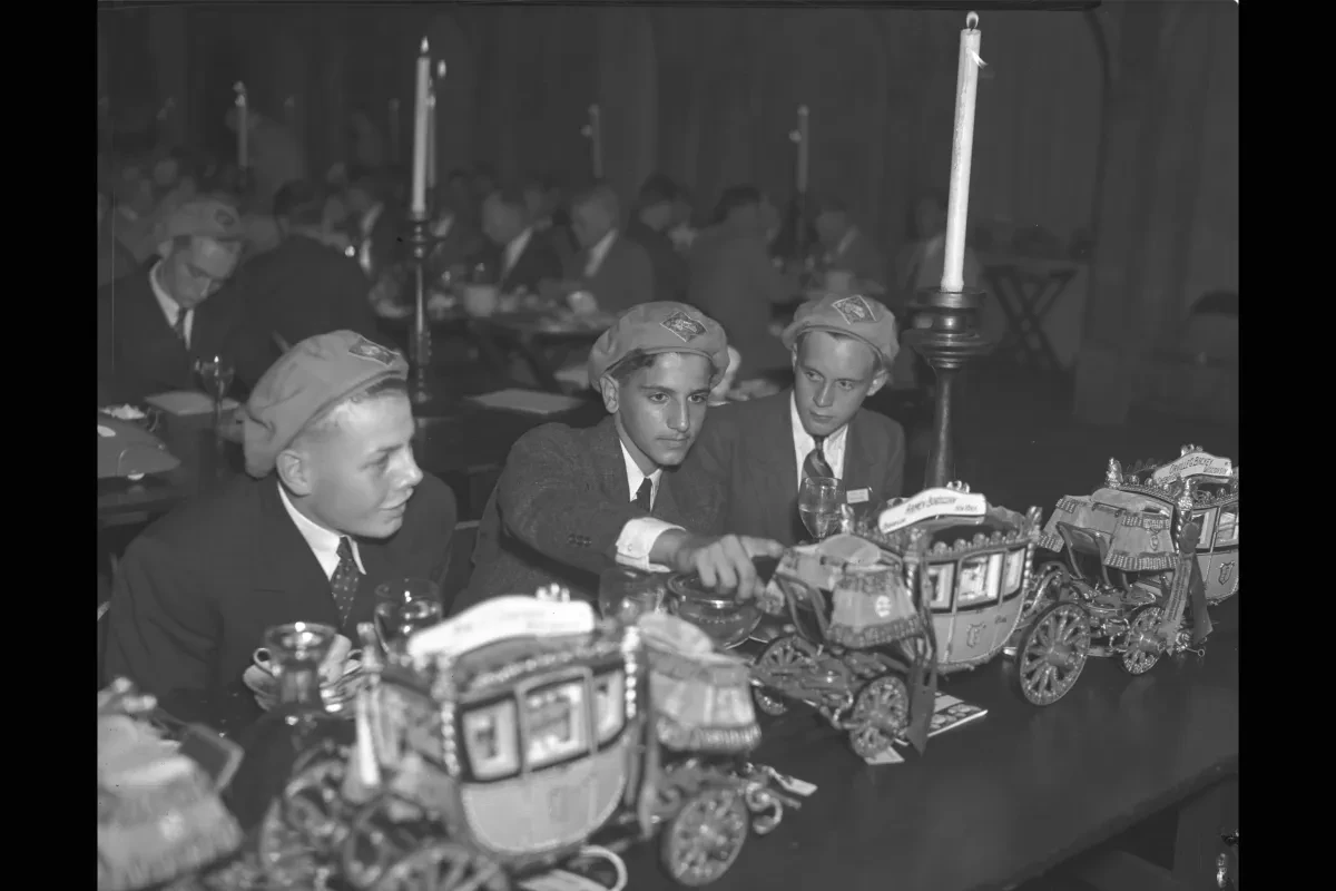 Three men in hats examine model stagecoaches on a table in a dimly lit room.