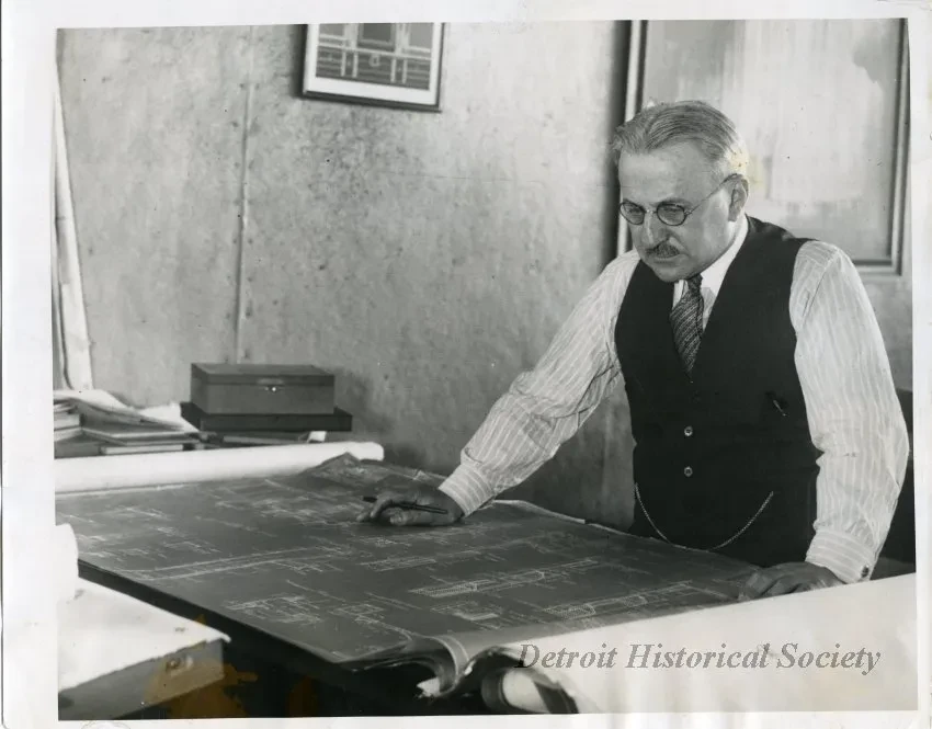 Man in vest and tie examines blueprints on a table in an office; appears focused and thoughtful.
