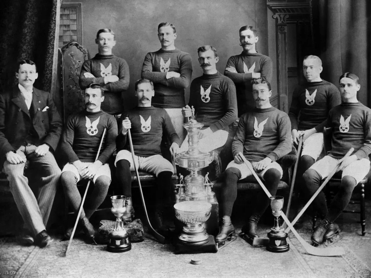 Vintage hockey team in dark uniforms poses with trophies, holding hockey sticks.