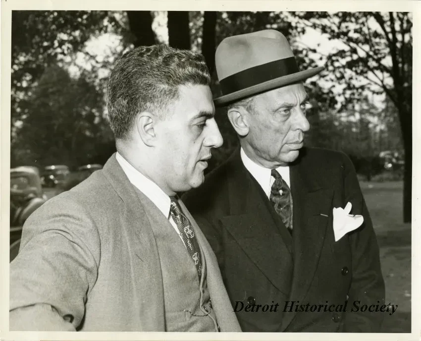 Two men in suits conversing outdoors, one wearing a hat. Trees and cars in the background. Black and white photograph.