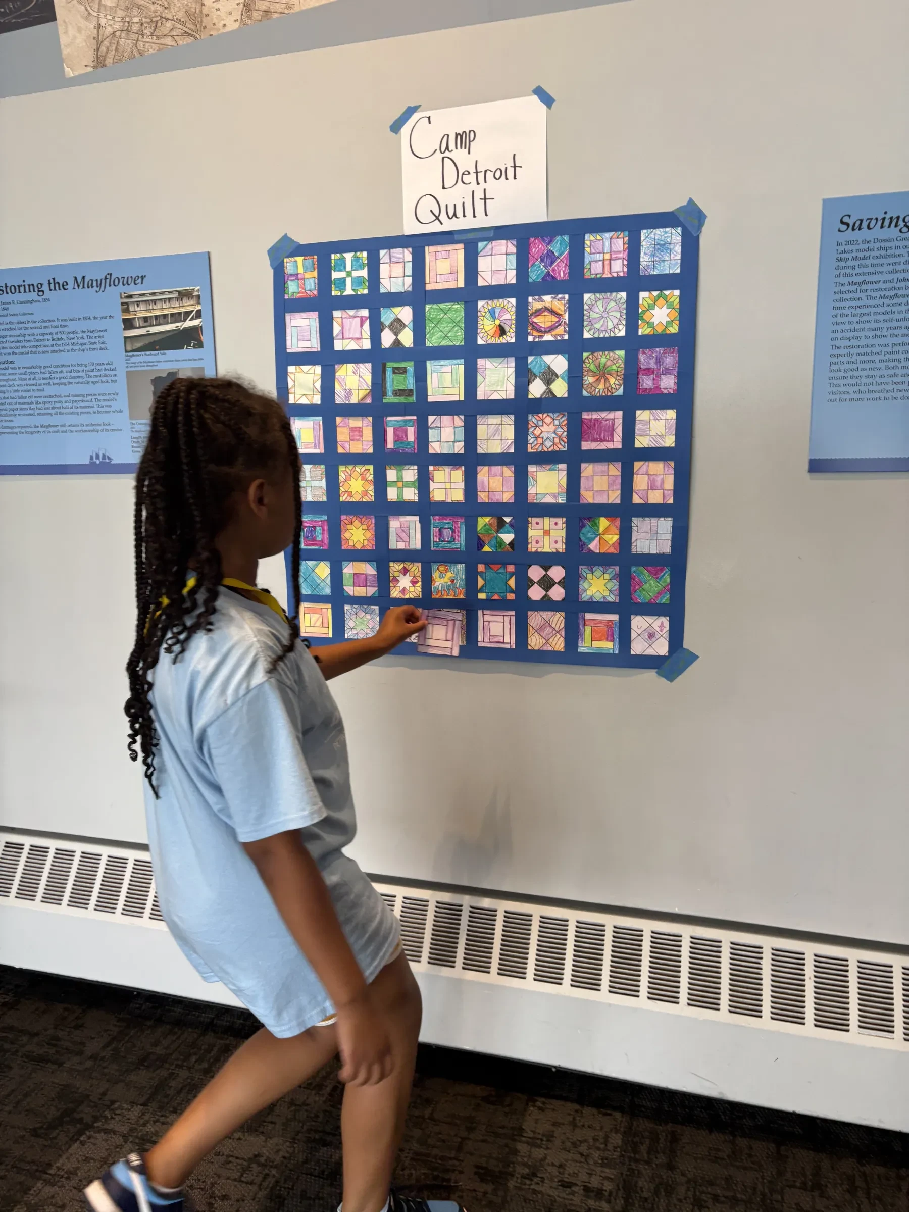 Child in blue outfit looks at colorful quilt displayed on a wall.