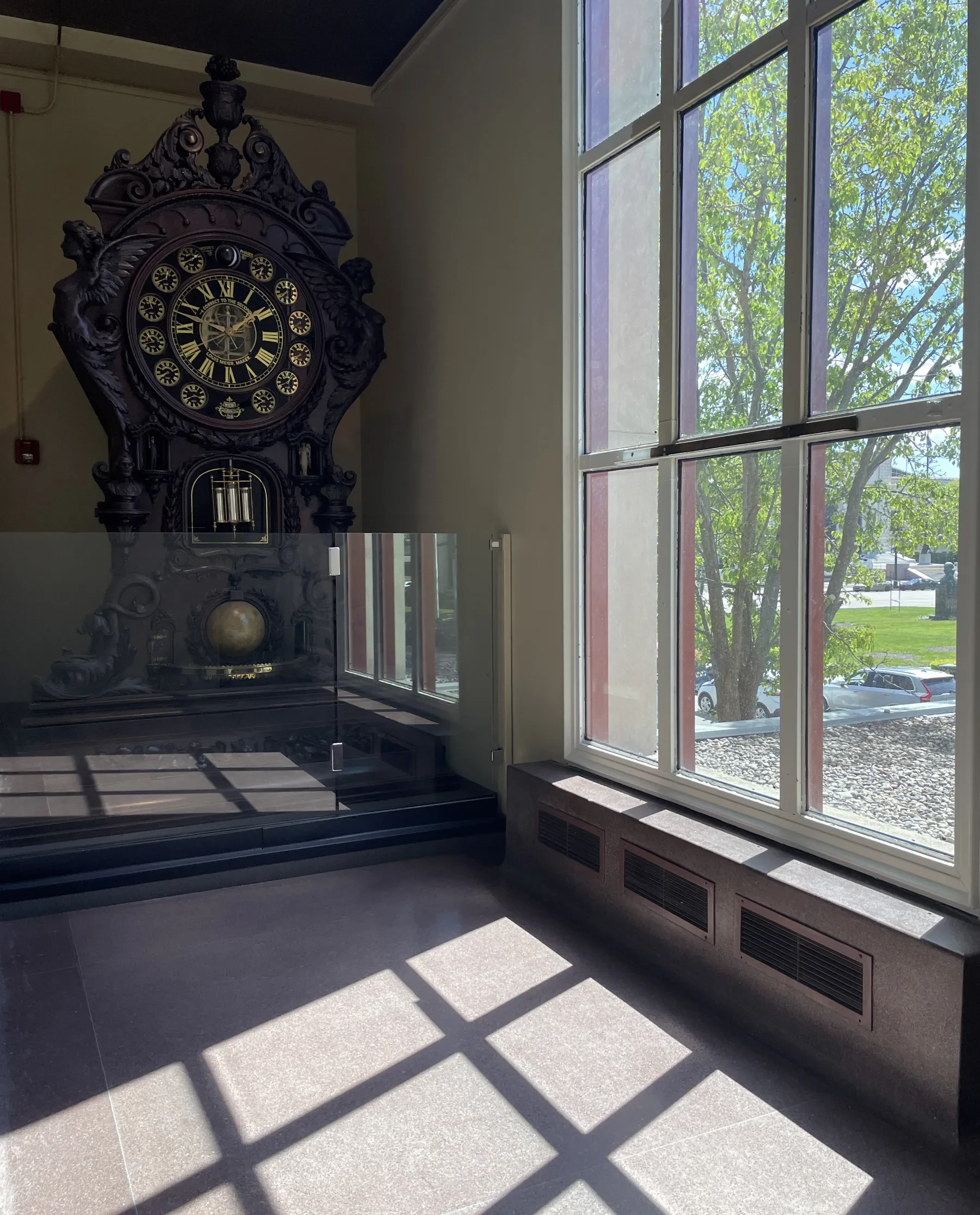 Ornate clock inside a room with large sunlit windows casting grid-like shadows on the floor.