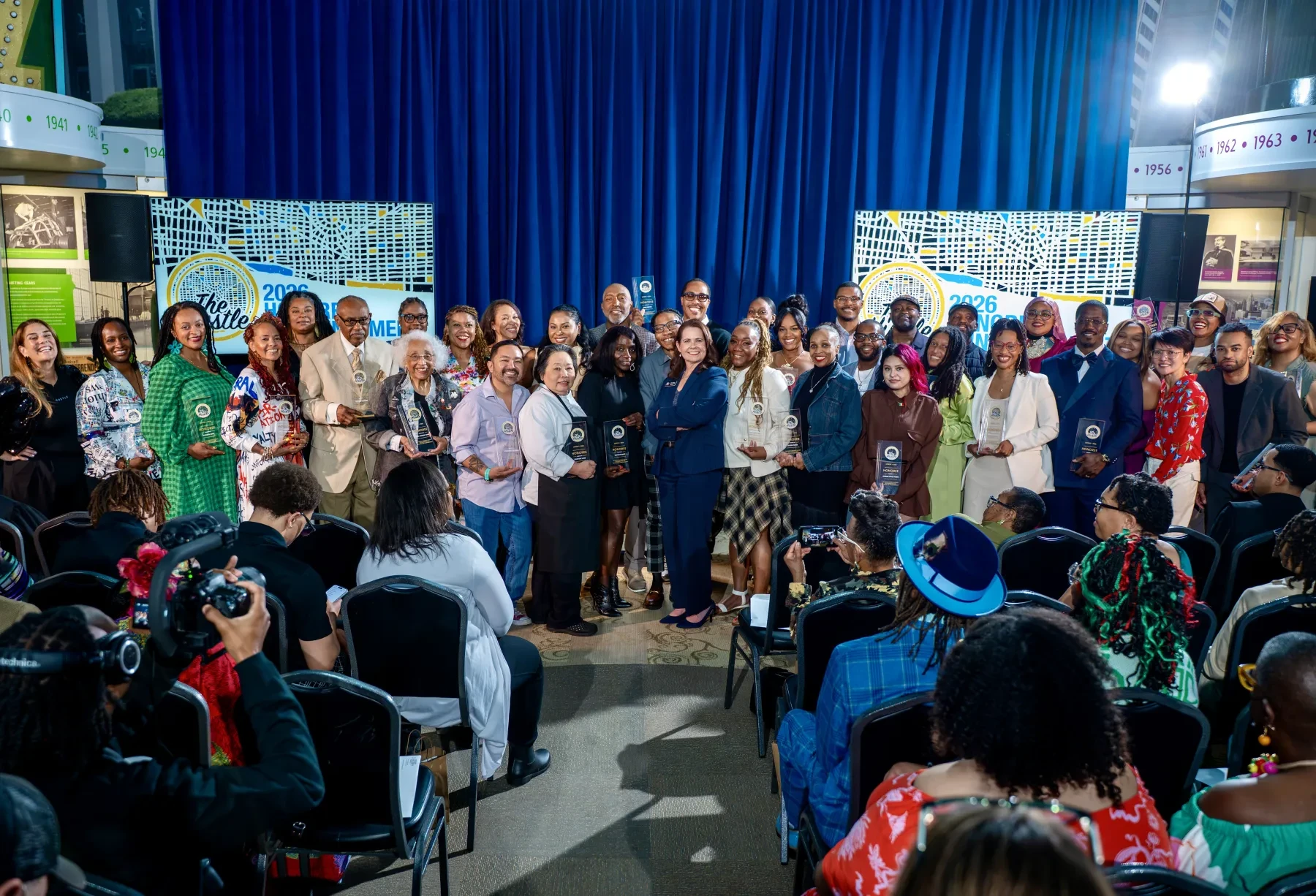 A large group of people stands on a stage facing an audience, with a blue curtain backdrop.