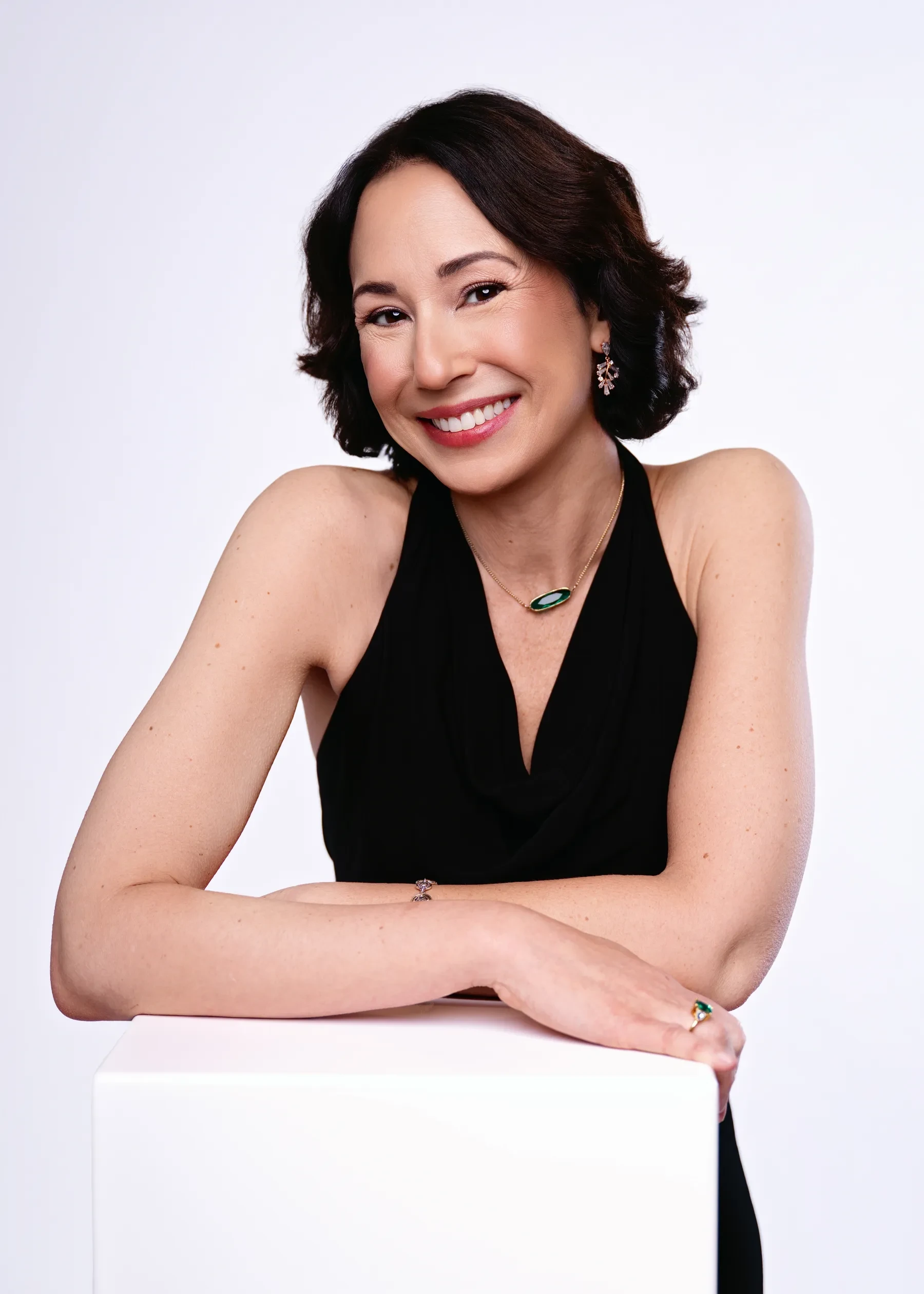 Smiling woman in black dress leaning on a white cube, against a plain white background.